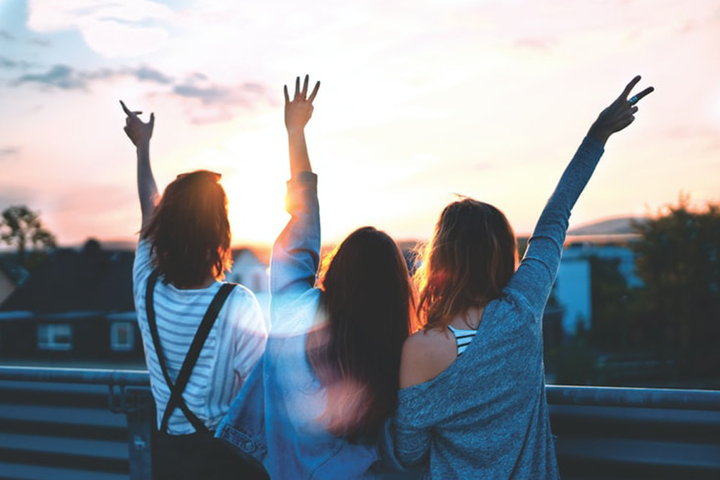 Drei jugendliche Frauen halten die Hände in die Luft und schauen sich den Sonnenuntergang über der Stadt an