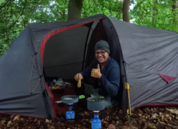 Zelt im Wald, Max schaut aus Zelt und sitzt kochend auf dem Boden