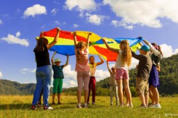 Kinder stehen auf einer Wiese im Kreis und halten ein buntes Tuch in die Luft