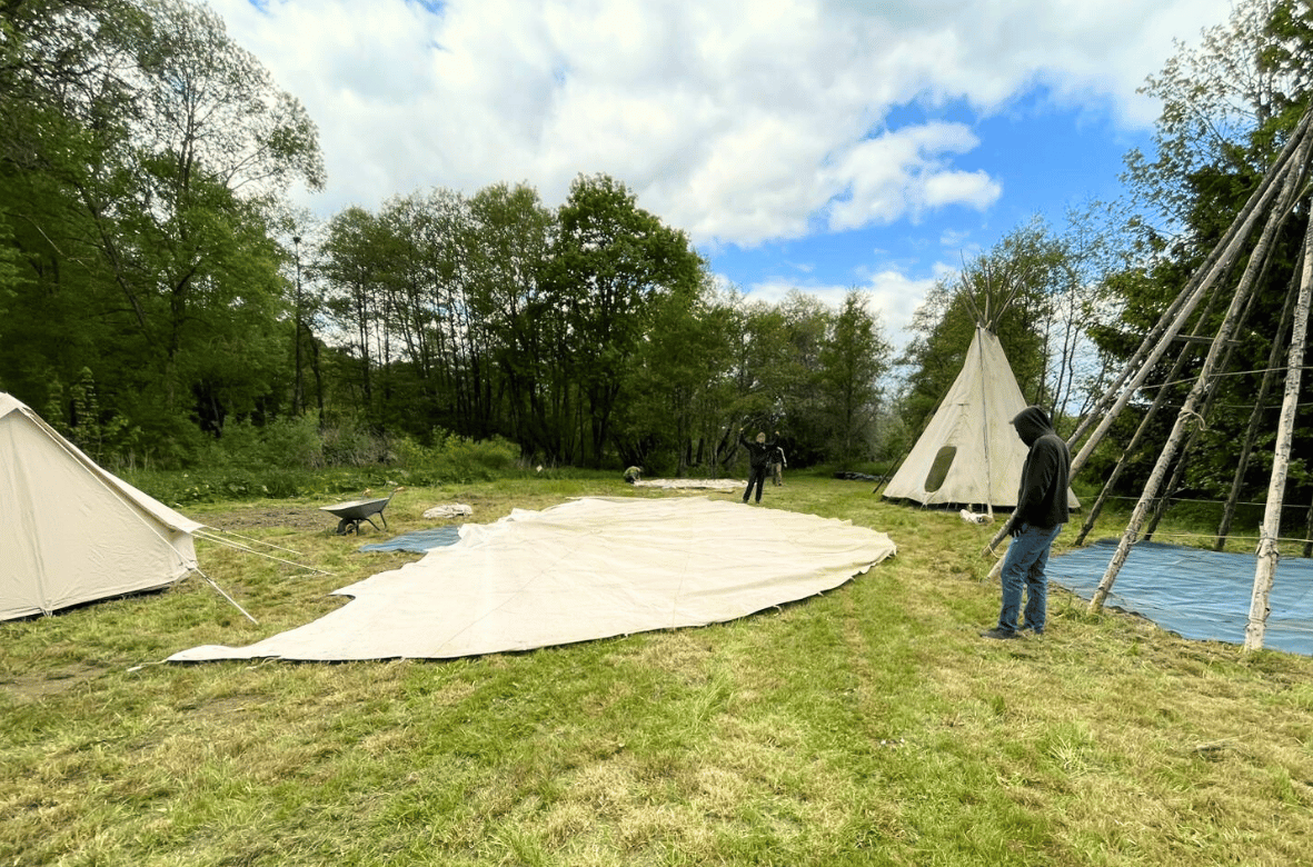 Menschen bauen das Tipi Dorf auf, auf dem Boden liegt eine Plane, einige Zelte sind schon fertig aufgebaut